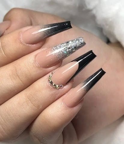 Mão com unhas longas estilo caixão, degradê nude e preto com glitter prata e pedrarias douradas